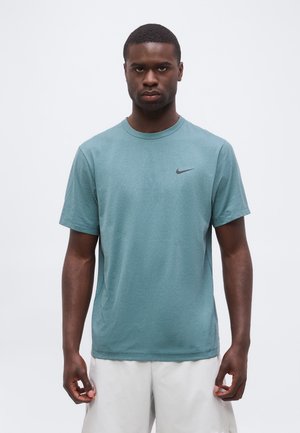 Mees kannab türkiissinist Nike lühikeste varrukatega särki ja valgeid lühikesi pükse, seisab neutraalse ilmega ühevärvilise tausta ees.