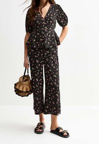 Top peplum negro con estampado floral y mangas abullonadas, combinado con pantalones a juego. La modelo lleva un bolso de paja y sandalias negras de slide.