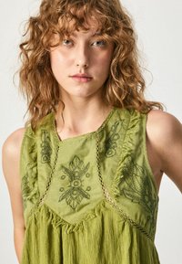Abito senza maniche verde ricamato con scollo sagomato, motivi floreali intricati e dettagli frangiati sull'orlo e sui lati.