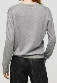 Pull gris en tricot avec manches longues et encolure ronde, porté avec un pantalon en denim noir. Coutures visibles et coupe décontractée.