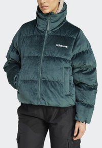 Grön sammets-puffjacka med hög krage, horisontell quiltad design, dragkedjestängning och broderad logotyp på vänster bröst.