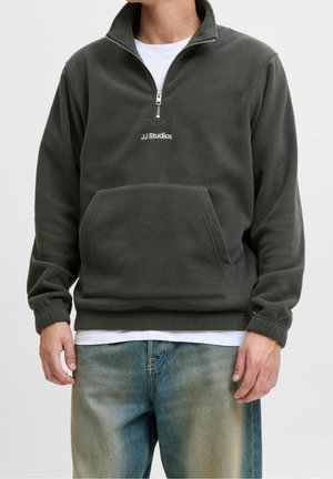 Homme portant une polaire vert foncé à fermeture éclair quart de zip avec poche avant et logo blanc "JJ Studios", associé à un jean bleu délavé et un t-shirt blanc en dessous.