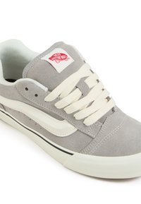 Sneaker in suede grigia con suola in gomma bianca, lacci crema e linea di accento nera. Presenta un'etichetta con il logo Vans sulla linguetta e la caratteristica striscia laterale.