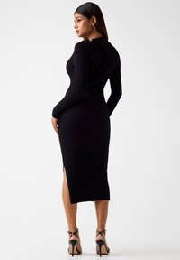 Czarna dzianinowa sukienka z długimi rękawami, o prążkowanej fakturze, dopasowanym kroju i rozcięciem z boku. Posiada kołnierzyk z ozdobnym guzikiem.