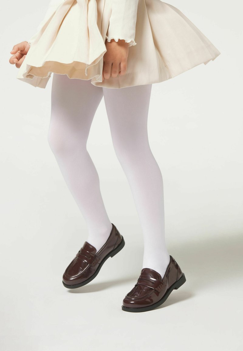 Braune Lacklederschuhe mit einer niedrigen schwarzen Sohle, getragen mit weißen Strumpfhosen unter einem beigen Kleid, das einen ausladenden Rock und gerippte lange Ärmel hat.