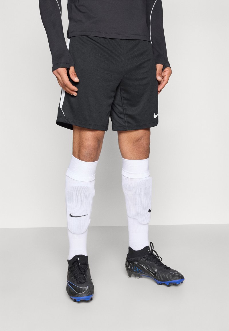 Schwarze Sportshorts mit weißen Seitenstreifen, kombiniert mit knielangen weißen Socken und schwarzen Fußballschuhen mit blauen Akzenten. Nike-Logo sichtbar.