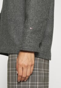 Cappotto in lana grigia con una vestibilità strutturata, caratterizzato da un polsino con bottone e una piccola toppa con logo sulla manica. Tessuto a quadri sotto.