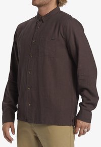 Brown, long-sleeve button-up shirt with a pocket, made from textured fabric. Features round buttons and a subtle brand label near the pocket.