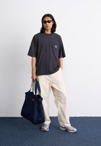 T-shirt oversized gris foncé avec poche poitrine, pantalon large beige, baskets argentées et sac fourre-tout bleu marine à pois avec poignées.