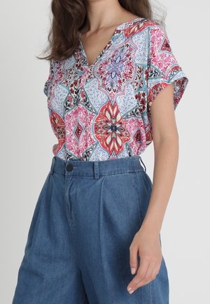 Femme portant un chemisier à manches courtes coloré et à motifs, rentré dans un pantalon ample en jean bleu taille haute, sur un fond uni.