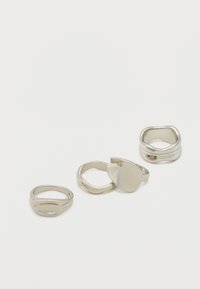 Quatre bagues en argent aux formes lisses et irrégulières, dont une avec un dessus plat ovale, disposées sur un fond blanc.