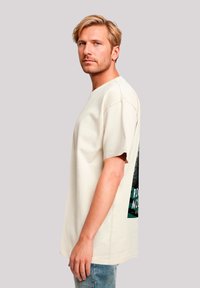 Blond man with beard stands sideways wearing a cream oversized t-shirt with printed graphic and light blue jeans against plain background.