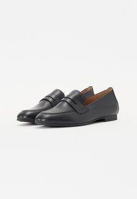 Mocassins en cuir noir avec une semelle plate, un bout arrondi et un design élégant avec une sangle décorative sur le devant. Intérieur doublé de marron.