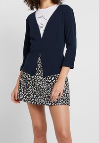 Navy blue fitted blazer with a V-neck, paired with a black skirt featuring a white leopard print pattern, showing 3/4 length sleeves.