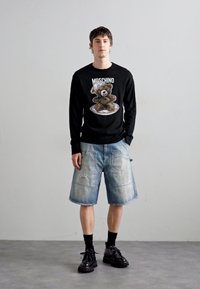 Cămașă neagră cu mânecă lungă, decorată cu un grafic de urs, asortată cu șorturi din denim bleu deschis. Pantofi negri cu talpă groasă și șosete închise completează ținuta.