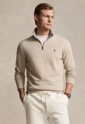 Uomo biondo con maglione beige con zip al collo e pantaloni bianchi, in piedi con le mani nelle tasche, guarda verso destra su uno sfondo neutro.