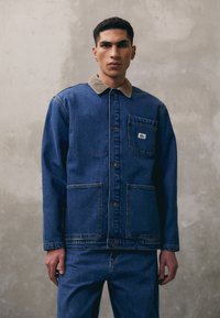Denimjacket met een beige corduroy kraag, voorzakken en knoopsluiting. Gedragen over bijpassende denim broeken. Glad textuur, blauwe kleur.