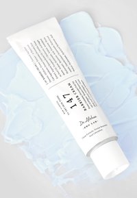 Hvit tube med barrierekrem med matt finish. Har svart tekst, en teksturert kork, og en illustrasjon av lys blå krem i bakgrunnen.