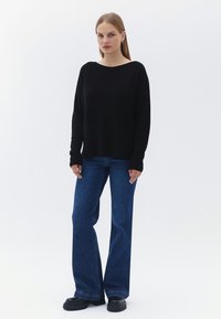 Černý pletený svetr s širokým výstřihem, spárovaný s modrými zvonovými džíny a černými platformovými botami. Modelka stojí na neutrálním pozadí.