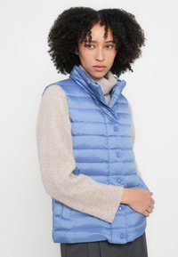 Jasnoniebieska pikowana kamizelka z wysokim kołnierzem, zapinana na guziki, o gładkiej fakturze, noszona na taupe swetrze.