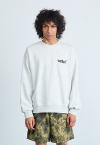 Heled halli sweatshirt ümmarguse kaeluse, soonikkoes varrukate ja musta logoga rinnal, kombineeritud roheliste kamoflaaziga lühikeste pükstega, millel on pikseliseeritud muster.