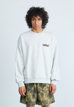 Heled halli sweatshirt ümmarguse kaeluse, soonikkoes varrukate ja musta logoga rinnal, kombineeritud roheliste kamoflaaziga lühikeste pükstega, millel on pikseliseeritud muster.