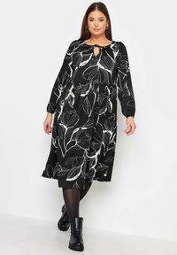 Mustat mekko, jossa on valkoisia lehtikuvioita, pitkät hihat ja avainaukko kaula-aukko. Viritetty muotoilu, jossa on kerätty vyötärö, yhdistettynä mustiin saappaisiin.