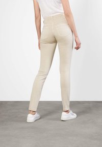 Beżowe, wysokim stanem, obcisłe jeansy z tylnymi kieszeniami, gładką fakturą i wąskim krojem, zestawione z białymi trampkami na szarym tle.