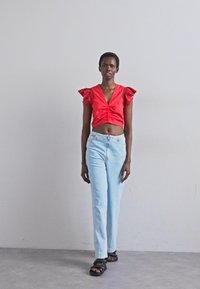 Rød kort topp med bølgede ermer, paret med lyseblå høyde i livet jeans. Svarte sandaler fullfører antrekket. Enkel bakgrunn.