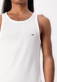 Person mit mittlerem Hautton, der ein weißes, geripptes Trägershirt mit einem kleinen roten, weißen und blauen Logo auf der Brust trägt, vor einem einfarbigen Hintergrund.
