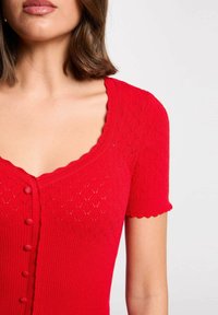 Il top lavorato a maglia rosso presenta maniche corte, un collo a scallop e una chiusura a bottoni. Ha un motivo a diamante testurizzato su tutto il capo.