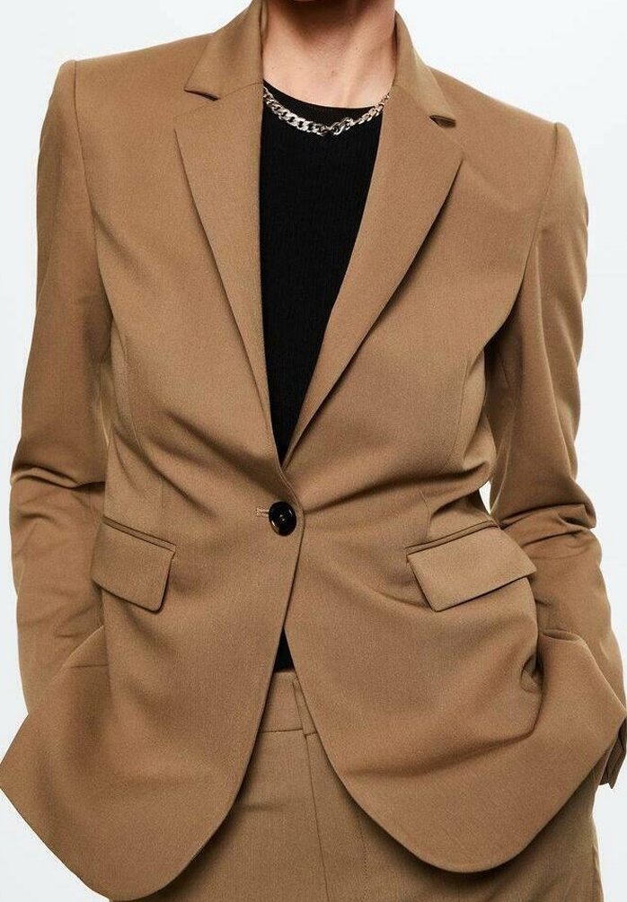 Personne portant un blazer beige à un bouton et un pantalon assorti avec un haut noir et un collier chaîne argenté. Mains dans les poches du blazer.