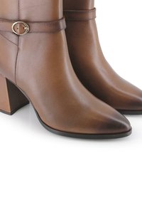 Botas de tobillo de cuero marrón con punta afilada y tacón grueso, que presentan una sutil correa con un detalle de hebilla circular. Textura suave.