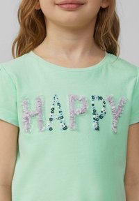 Lichtgroene katoenen t-shirt met het woord "HAPPY" versierd met meerkleurige bloemen-applicaties en pailletten. Korte mouwen, ronde hals.