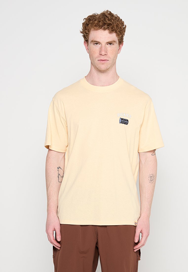 Uomo dai capelli rossi che indossa una t-shirt Dickies giallo pallido e pantaloni marroni, in piedi davanti a uno sfondo bianco semplice.