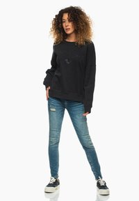 Svart sweatshirt med "Malibu" i rhinestones, ihop med slitna blå jeans och svarta plattformssneakers med vita sulor. Lockigt hår.