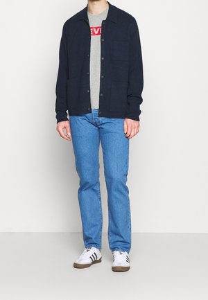 Marineblå cardigan over grå t-shirt med rød tekst, blå jeans og hvide sneakers med sorte striber. Har knapper og lommer foran.
