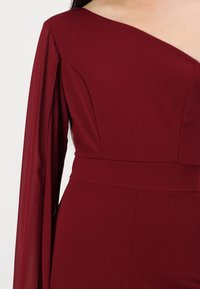 Vestido burdeos con escote asimétrico, manga larga transparente y corsé ajustado. Textura suave, silueta entallada y detalles de costura sutiles.