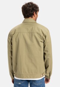 Khaki jas met een kraag, knoopsluitingen aan de mouw en zijknopen. Heeft een ontspannen pasvorm en een gladde, lichte stof.