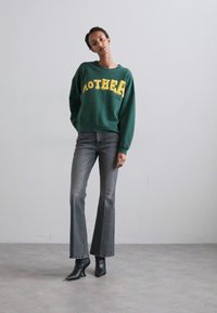 Felpa verde con testo giallo che dice "MAMMA". Abbinata a jeans grigi svasati e stivali neri con tacco. Tessuto morbido con vestibilità rilassata.