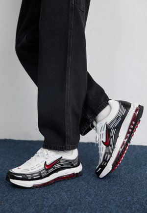 Personne portant un pantalon noir et des baskets Nike blanches, noires et rouges, debout sur un tapis bleu.