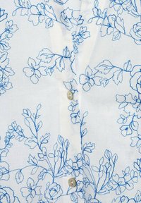 Camicia di tessuto bianco con ricami floreali blu, dotata di chiusura con bottoni sul davanti. Design floreale semplice con dettagli di cucitura ben visibili.