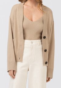 Kvinna iklädd en beige ribbad tanktop, matchande kardigan med svarta knappar, och hög midja i off-white byxor.