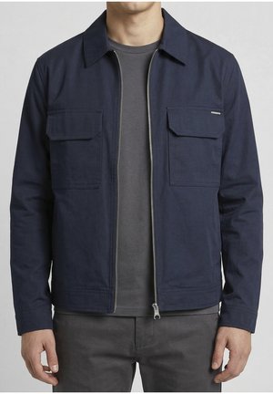 Hombre con una chaqueta azul marino con cremallera y bolsillos con solapa en el pecho, sobre una camiseta gris oscura y pantalones grises, de pie contra un fondo liso.