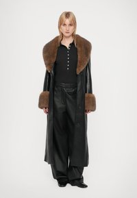 Donna bionda in piedi, che indossa un lungo cappotto in pelle nera con collare e polsini in pelliccia marrone, una maglietta nera, pantaloni larghi in pelle nera e scarpe nere.
