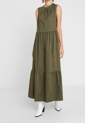 Oliwkowa, bezrękawnikowa sukienka maxi z marszczonymi warstwami, noszona z białymi botkami na obcasie, modelka stoi na tle gładkiego, jasnego tła.