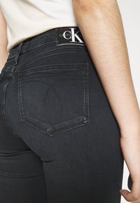 Jeans de mezclilla oscura con cintura alta, con una etiqueta de Calvin Klein en la parte trasera. La tela tiene una textura suave y sutiles detalles de lavado.