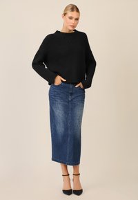 Schwarzer gestrickter, verkürzter Pullover mit Rippstruktur, kombiniert mit einem langen Jeansrock mit leichten Verzierungen und schwarzen High Heels.