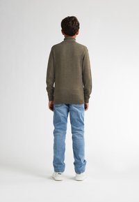 Maglione verde scuro con colletto alto, abbinato a jeans azzurri chiari. L'outfit presenta una vestibilità rilassata, che mette in mostra texture morbide e dettagli minimi.