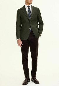 Dunkelgrüner Wollblazer mit Revers, kombiniert mit einem weißen Hemd und einer gestreiften marineblauen Krawatte; braune Cordhosen und dunkle Lederschuhe.